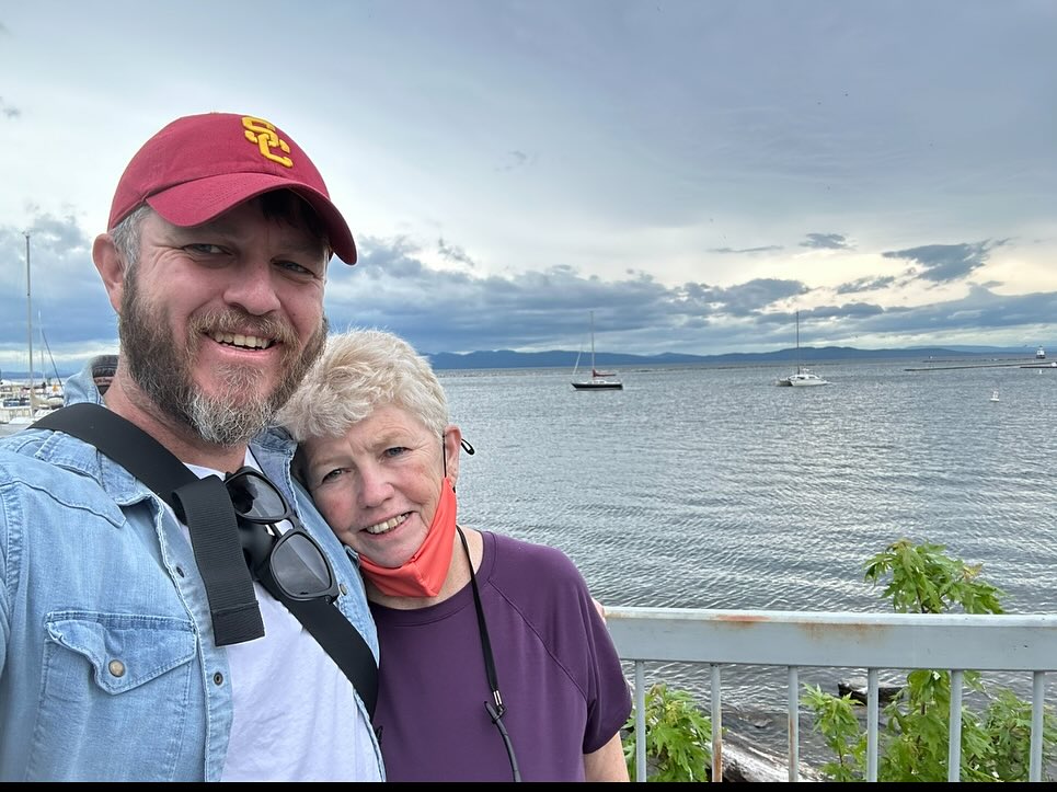 Chris and Joan smiling together at a harbor, sailboats on the water behind them