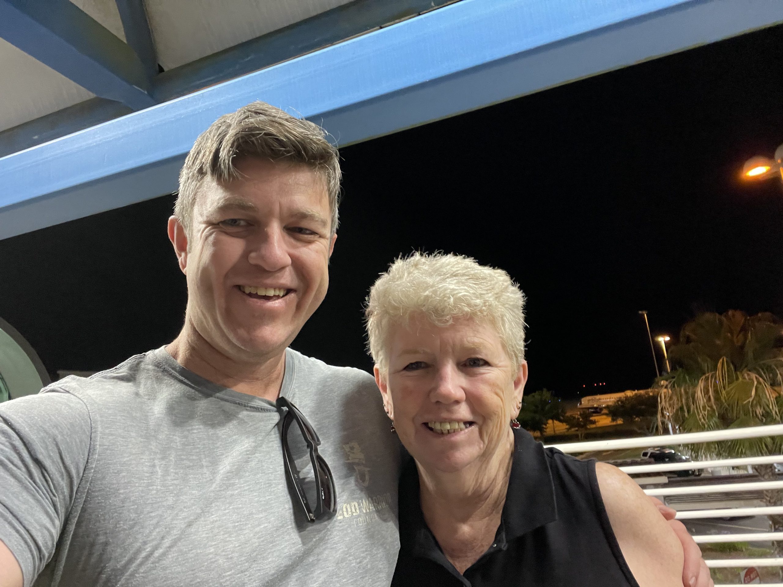 Chris stands beside his mother, Joan, both smiling warmly at the camera. Joan is wearing a light blue scarf and glasses, her arm gently around Chris. They appear close, comfortable, and full of love—a quiet, tender moment between mother and son.